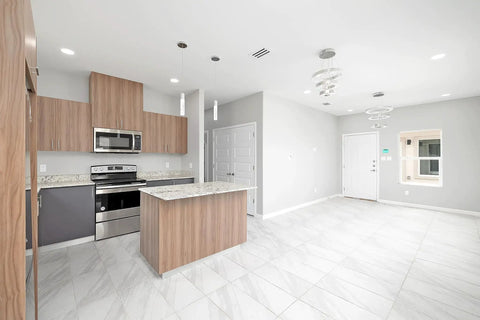 Modern kitchen with wooden cabinets and stainless steel appliances next to empty living room with white walls and tiled floor.