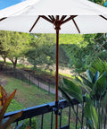 Adjustable Outdoor Umbrella Clamp Holder That Clips On Rails Benches Decks And More Universal Umbrella Clamp Mount Fits 0.8 to 2.7 Inch Railings Fences Balconies Tailgates Boats And Patio Spaces securing an umbrella to a balcony rail with plants and trees in the background.