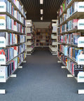 Library aisle with labeled book shelves; signs for Auxiliary Science, Anthropology, World History, and Literature sections visible.