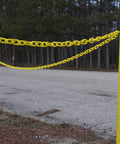 Two yellow poles with Yellow Plastic Barrier Chain With 2 Inch Links, 50 Foot Length For Crowd Control And Traffic Direction, Lightweight Yet Durable Barrier For Events And Worksites, Cut To Fit With Snips Or Scissors strung between them, outdoor setting.