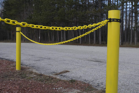 Two yellow poles with Yellow Plastic Barrier Chain With 2 Inch Links, 50 Foot Length For Crowd Control And Traffic Direction, Lightweight Yet Durable Barrier For Events And Worksites, Cut To Fit With Snips Or Scissors strung between them, outdoor setting.