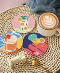 Three diamond-painted cocktail coasters on a wicker surface with a latte, part of the Eight Piece Diamond Art Coaster Kit With Holder For DIY 5D Retro Cocktail Coasters Diamond Painting Set For Adults And Beginners Includes Adhesive Cork Bases Diamond Tiles And Marker Pen.