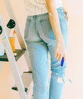 Person holding a heavy duty paint roller with frame, wearing jeans while on a ladder for easy wall painting.