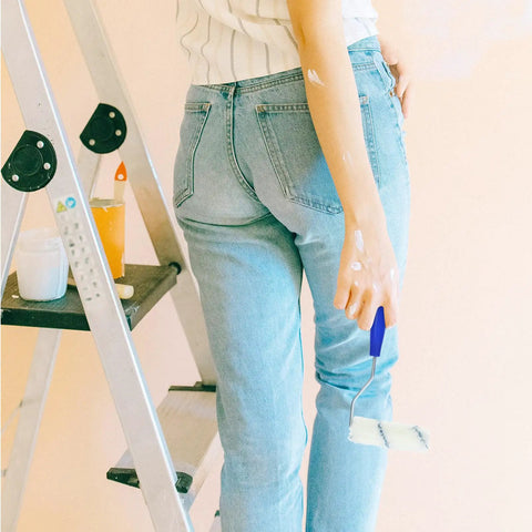 Person holding a heavy duty paint roller with frame, wearing jeans while on a ladder for easy wall painting.