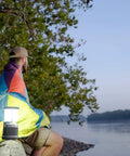 Man wrapped in blanket near river with 60 Day Portable LED Lantern With 1200 Lumen Power on wooden post at dusk.