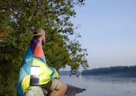 Man wrapped in blanket near river with 60 Day Portable LED Lantern With 1200 Lumen Power on wooden post at dusk.