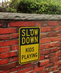 Heavy Duty Slow Down Kids Playing Neighborhood Warning Sign Crafted From Tough 63 Mil Aluminum on a brick wall outdoors.
