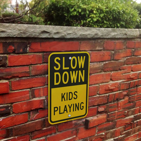 Heavy Duty Slow Down Kids Playing Neighborhood Warning Sign Crafted From Tough 63 Mil Aluminum on a brick wall outdoors.