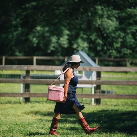 Woman walking outside carrying a pink soft sided cooler bag at a sunny park.