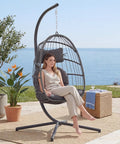 Woman sitting in Indoor Outdoor Egg Chair With Stand For Patio And Balcony Wicker Swing Egg Chair Rust Resistant Steel Frame And Heavy Duty Stand Adjustable Suspension Chain 350 Lb Capacity Charcoal Grey near the ocean.