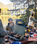 Group of friends camping by a lake, grilling food on a portable stove, picnic setup with tents and trees in the background.