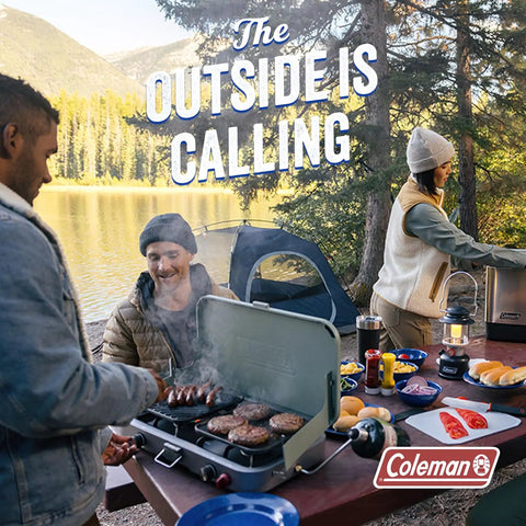 Group of friends camping by a lake, grilling food on a portable stove, picnic setup with tents and trees in the background.