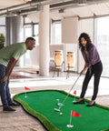 Two people using Pro Grade Golf Putting Green Practice Mat 5x10ft with flags and cups in an office setting.