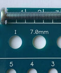 Close-up of blue-green Holistic drill bit gauge with three sizes: 17/64 in, 7.0 mm, 9/32 in; metal screw resting on top.
