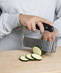 Stainless steel crinkle cutter with black handle, side view slicing cucumber on a wooden cutting board.