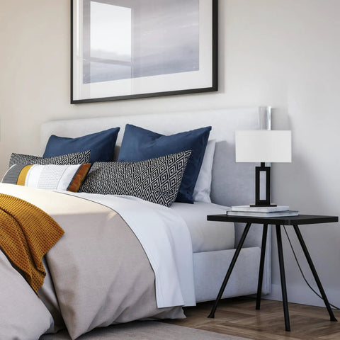 D'Alessio Table Lamp In Matte Black With Modern Cutout Body And White Linen Shade, 20 Inch Tall on a bedside table next to bed.