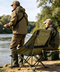 Two people fishing by a river with a foldable fishing chair and gear, outdoor setting with trees and water.