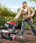 Person using a red and black garden tiller on soil with green background, wearing a beige jacket and jeans.