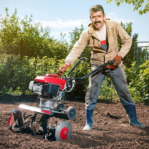Person using a red and black garden tiller on soil with green background, wearing a beige jacket and jeans.