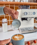 Hand pouring milk into a cup of coffee in front of a Semi Automatic Espresso Machine With Built In Burr Grinder And Smart Dosing Tech For Smooth One Or Two Shot Pulls Sturdy Metal Body In Porcelain White Delivers Legit Cafe Quality Straight Outta The Box.