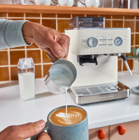 Hand pouring milk into a cup of coffee in front of a Semi Automatic Espresso Machine With Built In Burr Grinder And Smart Dosing Tech For Smooth One Or Two Shot Pulls Sturdy Metal Body In Porcelain White Delivers Legit Cafe Quality Straight Outta The Box.