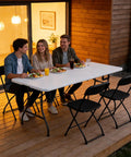 Three people sitting outdoors on a Six Foot Folding Table Portable Picnic Table For Indoor Outdoor Use Heavy Duty Plastic Table