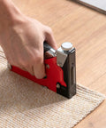 Hand using Beast Of A 4 In 1 Heavy Duty Upholstery Staple Gun Kit on carpet edge for woodworking and DIY furniture projects