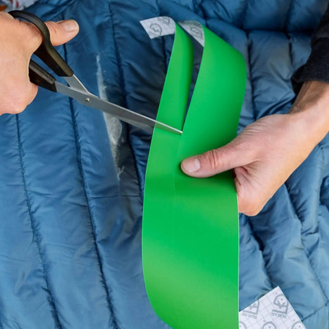 Person cutting Tenacious Tape 3 Inch By 20 Inch Clear Vinyl Gear Repair Tape With Two 1.5 Inch Hex Patches on a blue jacket
