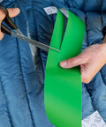 Hands cutting green repair tape with scissors on a blue jacket, demonstrating use of Tenacious Tape 3 Inch By 20 Inch Nylon Fabric Repair Tape Patch For Holes And Rips In Jackets Rain Gear Tents Sleeping Bags And Outdoor Gear Peel And Stick No Heat Or Sewing Required Black 1 Roll.