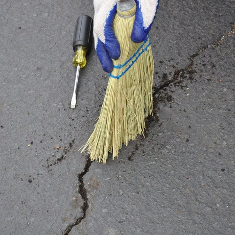 Person preparing asphalt crack for PLI-STIX 60 Ft Asphalt And Concrete Crack Filler 2 Pack For Driveways Self Leveling And Watertight Seal Pro Grade Crack Repair That Works With A Propane Torch Or Heat Gun application with a broom and screwdriver.