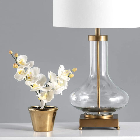 Hendry 23 Inch Glass Table Lamp with Brass Finish, Fabric Drum Shade, Inline Switch next to a potted orchid on a table