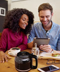 Man and woman at table with a 12 Cup Drip Coffee Maker With Auto Off And Reusable Filter, Borosilicate Glass Carafe, Anti Drip Water Window in black.