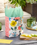 Nine Inch Medium Gift Bag With Tissue Paper Bright Flower Designs For Valentine Day Easter Birthdays Bridal Showers Weddings Baby Showers And Mothers Day A Cute Floral Gift Bag For Any Special Occasion on a patio table.