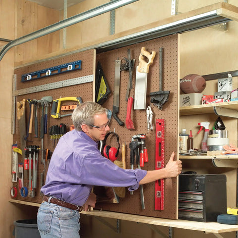Man organizing tools on a pegboard in a workshop, standing beside shelves filled with various items.