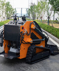 Compact orange tracked mini skid steer loader with backhoe attachment on urban street; hydraulic lifting and control panel visible.