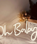 Oh Baby Neon Sign White LED Wall Decor With Dimmable Brightness And Plug In Power Adapter, illuminated on a wall near flowers.