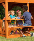 Children sitting at the picnic table of the Skyfort II Cedar Wood Swing Set With Wave Slide, Raised Clubhouse, Crow's Nest.