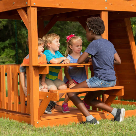 Children sitting at the picnic table of the Skyfort II Cedar Wood Swing Set With Wave Slide, Raised Clubhouse, Crow's Nest.