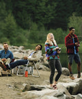 Four people enjoying fishing and relaxing by a lakeside campfire, holding fishing rods and reel combos.