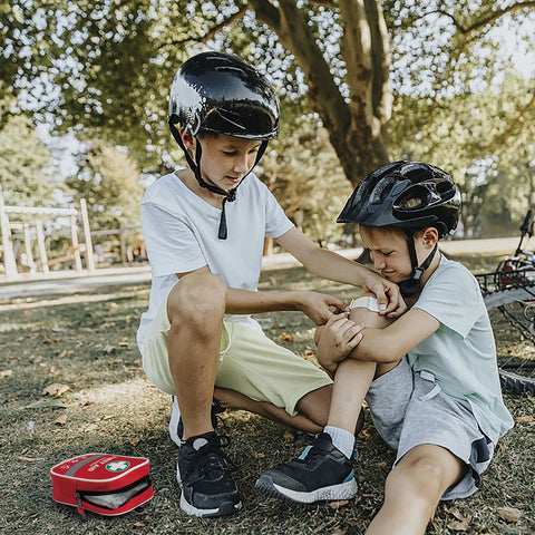Red first aid kit on grass beside two kids in bike helmets; one bandages the other's knee.
