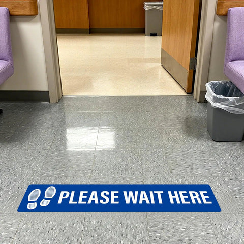 Five Piece Please Wait Here Floor Sign Kit 26 By 6 Inches Self Adhesive Waterproof Vinyl, Durable Social Distancing Markers For Queues And Waiting Areas, Multipack Signage For Busy Shops. Blue floor sign on hallway tile.