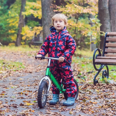 Child in star-patterned outfit holding green balance bike on leaf-covered path in autumn forest next to a bench.