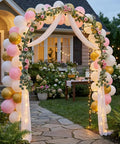 Balloon-decorated garden arch at outdoor event with flowers, pathway, and house backdrop; balloons in pink, white, and gold.