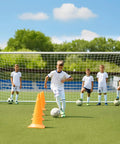 Children practicing soccer drills in front of 12x6 Ft Foldable Steel Backyard Soccer Goal With Weatherproof Net Perfect For Teens And Adults Practice Drills Easy Setup Portable Training Goal Set Includes 6 Training Cones.