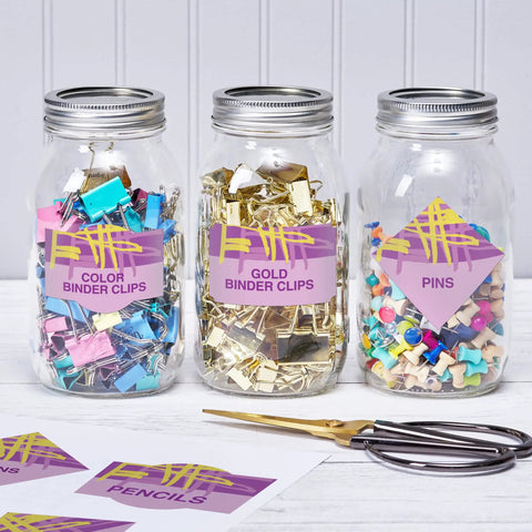 Three glass jars filled with colorful binder clips and pins, labeled Color Binder Clips, Gold Binder Clips, and Pins.