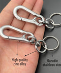Hand holding two keychain clips, one labeled high quality zinc alloy, the other durable stainless steel, against a dark background.