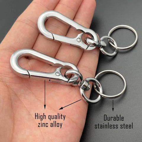 Hand holding two keychain clips, one labeled high quality zinc alloy, the other durable stainless steel, against a dark background.