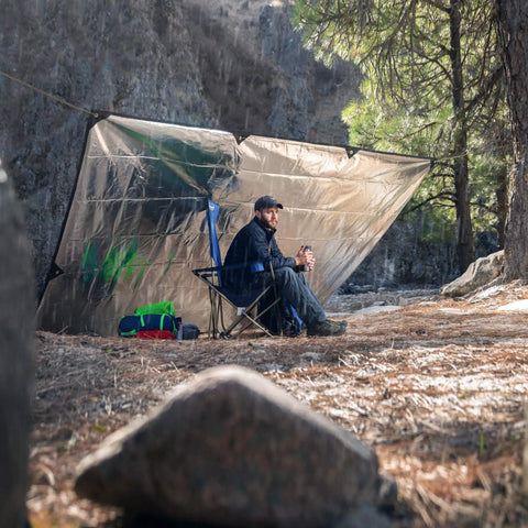Camper sitting beside Tough As Nails Heavy Duty Survival Blanket With Reflective Thermal Layer For Camping And Outdoor Gear Waterproof Tarp.