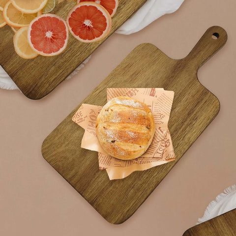 Acacia wood cutting board with handle, top-down view; bread roll on parchment