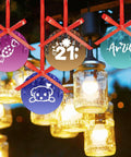 Colorful hanging ornaments with red ribbons, candy, bear, and text designs, glowing lights in background.